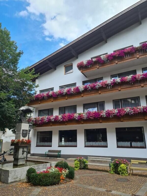 A multi-story building with balconies adorned with flowers, a paved courtyard, and a bench.