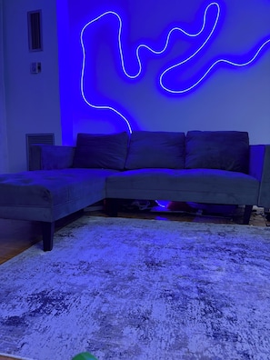 A modern living room with a sectional sofa, a patterned rug, and a wall with a blue neon light design.