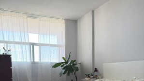 Un dormitorio con una cama, una ventana con cortinas traslúcidas, una planta en maceta y un aparador.