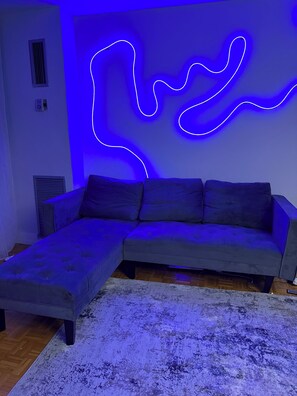 A modern living room with a grey sectional sofa, a white textured rug, and a blue neon light installation on the wall.