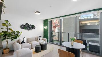 A modern living room with a sofa, coffee table, and chairs, a balcony with a view of buildings and a crane, and a green wall decoration.