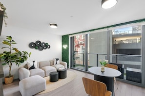 Ein modernes Wohnzimmer mit einer Couch, einem Couchtisch und Sesseln, ein Balkon mit Blick auf Gebäude und einen Kran, sowie eine grüne Wanddekoration.