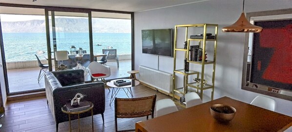 Ein modernes Wohnzimmer mit einem großen Fenster mit Blick auf ein Gewässer, einem Fernseher und einem goldrahmigen Bücherregal.