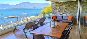 Ein Balkon mit Holztisch und -stühlen, einer Steinmauer und Blick auf das Meer und die Berge.