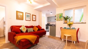 A living room with a red sofa, a dining table with chairs, a kitchen in the background, and framed pictures on the wall. - Cosy 2 Bed Viking Bay Cottage (Broadstairs)