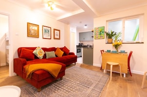 A living room with a red sofa, a dining table with chairs, a kitchen in the background, and framed pictures on the wall.