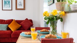 A dining table with plates, glasses, and croissants, a red sofa with cushions, and a vase with yellow flowers. - Cosy 2 Bed Viking Bay Cottage (Broadstairs)