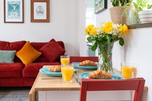 Una mesa de comedor con platos, vasos y croissants, un sofá rojo con cojines, y un jarrón con flores amarillas.