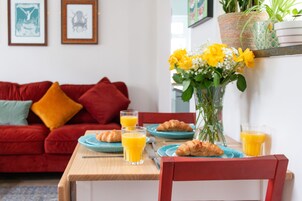 A dining table with plates, glasses, and croissants, a red sofa with cushions, and a vase with yellow flowers.