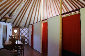 Ein Jurteninneres mit einem Holztisch und -stühlen, einer Tür mit rundem Fenster und einem roten Vorhang.