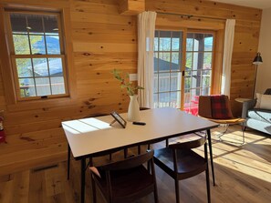 Un interior de madera con una mesa, sillas, un jarrón con flores y vistas a través de grandes ventanales.