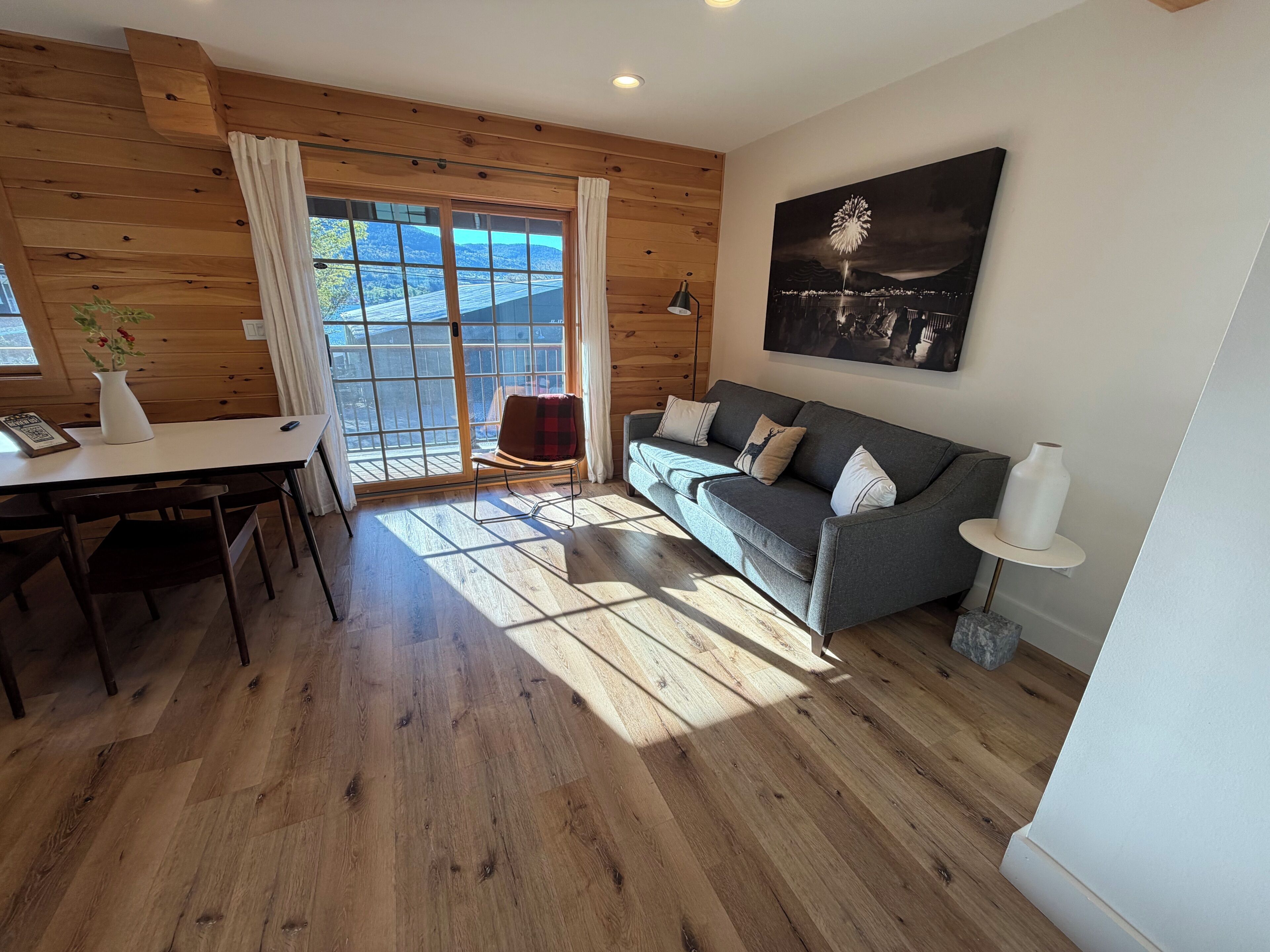 A modern living room with wooden walls, a grey sofa, a dining table with chairs, and a large window with a view of the outdoors.