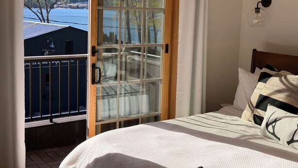 A bedroom with a bed, a window with a view of a lake and buildings, and a curtain. - Deluxe Lodge Lakefront On Lake George #11 (Lake George)
