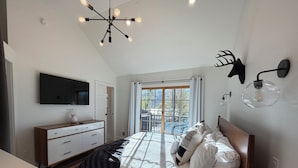 A bedroom with a bed, a TV, a dresser, a chandelier, and a window with a view. - Deluxe Lodge Lakefront On Lake George #11 (Lake George)