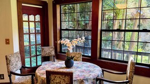 Un coin repas avec une table ronde, des chaises à motifs floraux, un vase rempli de fleurs et de grandes fenêtres donnant sur un paysage verdoyant.