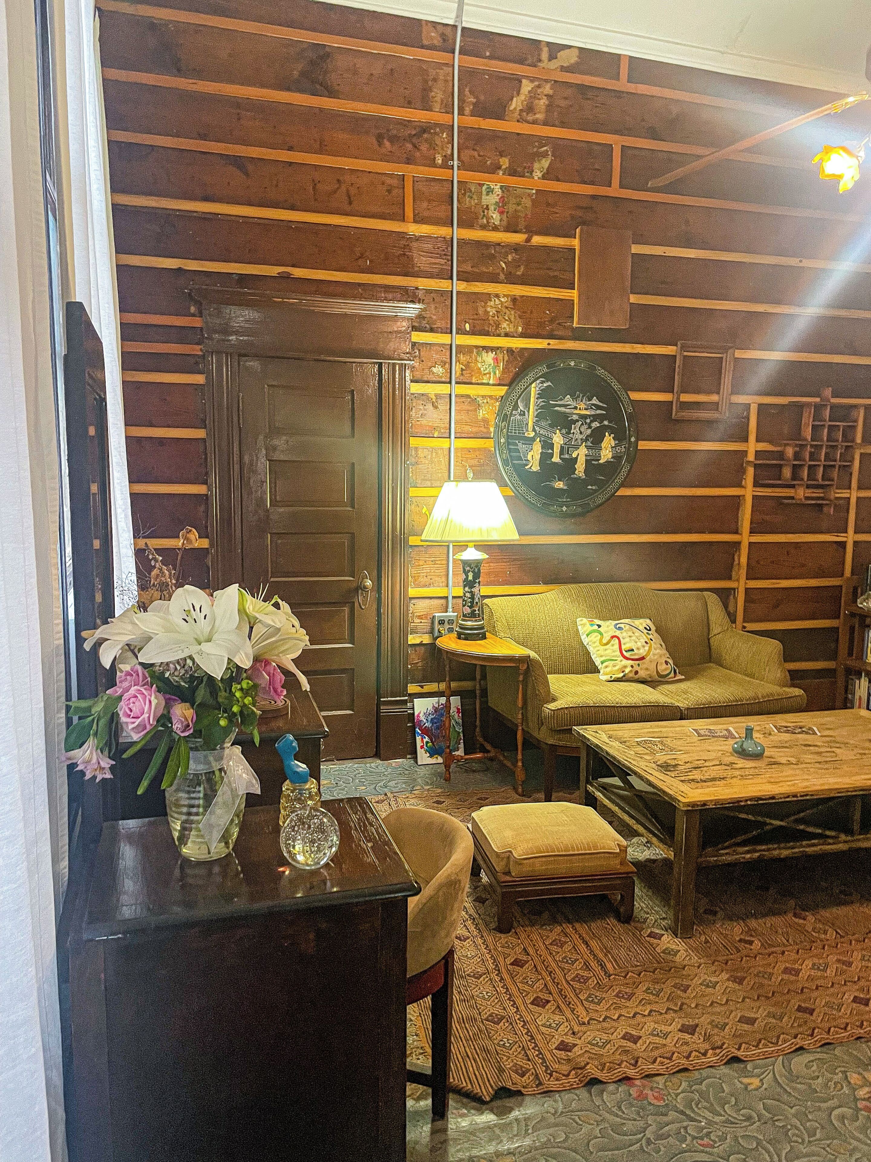 A room with a wooden wall, a sofa, a coffee table, a lamp, and a vase with flowers.