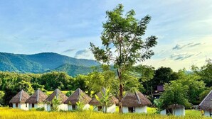 Cabañas de techo de paja en un campo con un árbol y montañas al fondo.