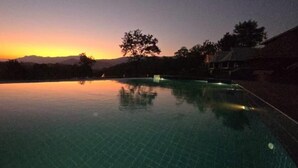 Una piscina al anochecer con vista a la puesta de sol, rodeada de árboles y un edificio.