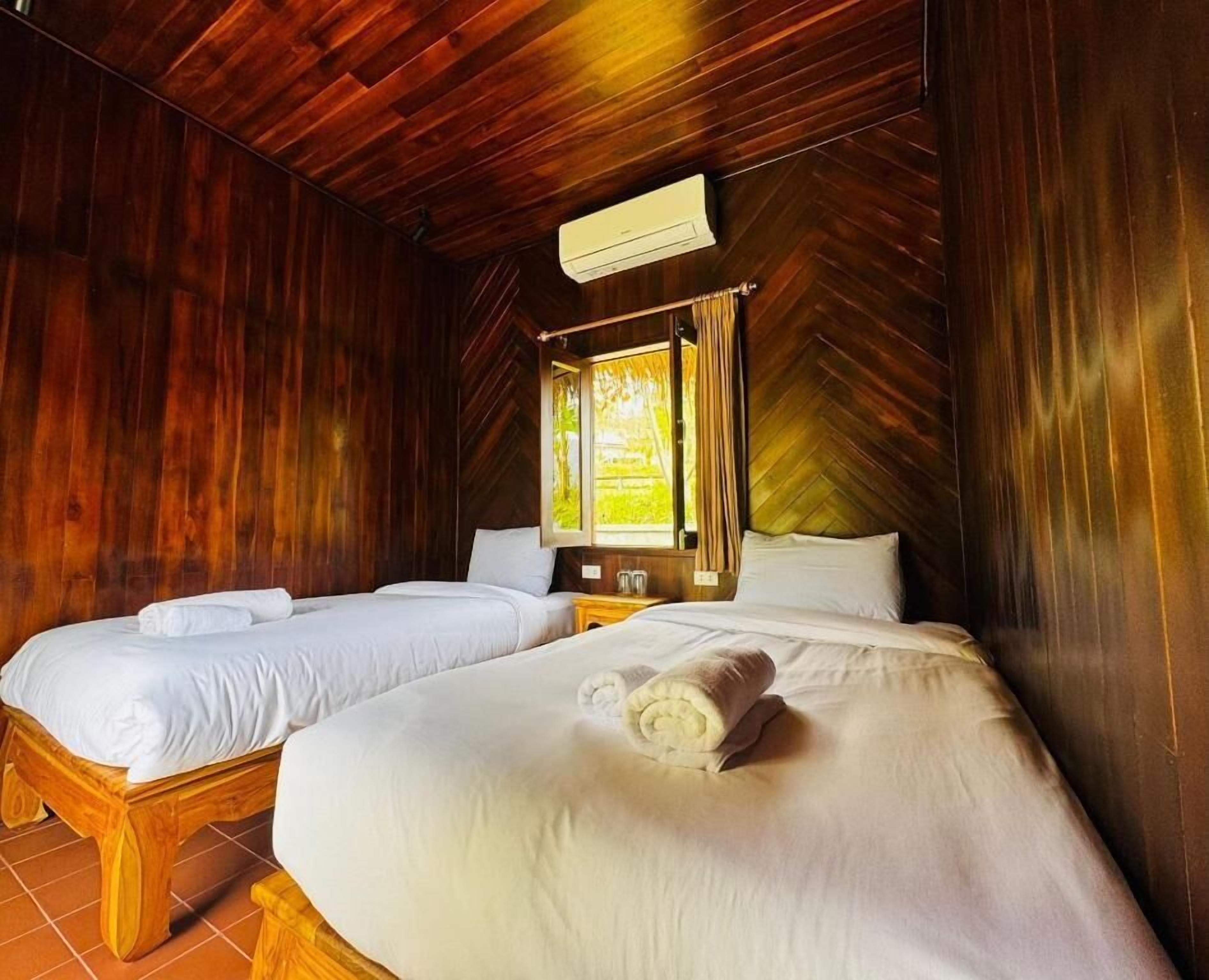 A room with two beds, wooden walls, a window with curtains, and an air conditioning unit.
