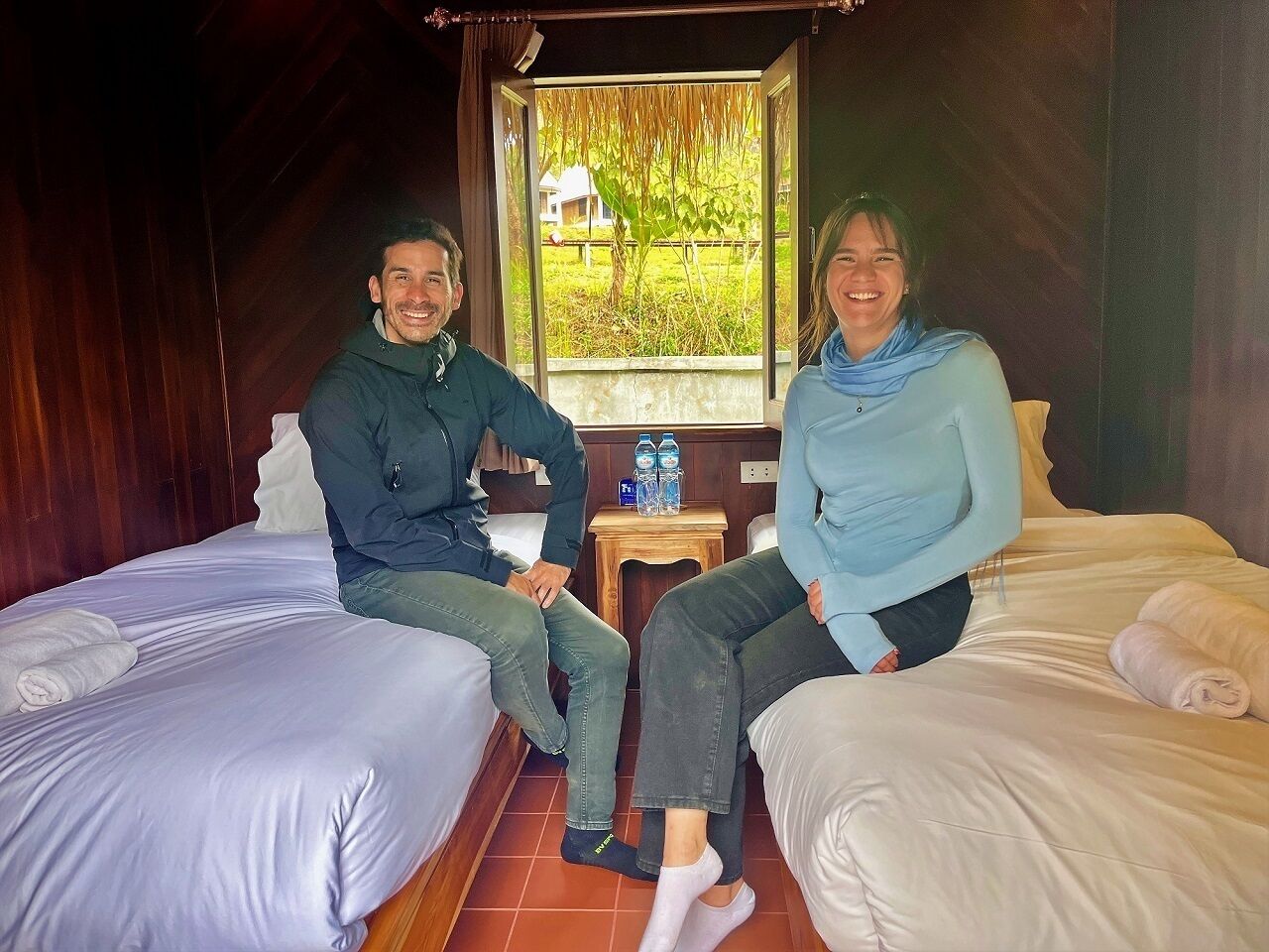 Ein Zimmer mit zwei Betten, einem Fenster mit Blick auf Grünflächen und einem kleinen Tisch mit Wasserflaschen.
