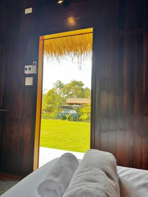 Ein Zimmer mit einem Bett, einem Fenster mit Blick auf eine grüne Wiese und ein Gebäude sowie einer Tür mit strohähnlicher Struktur.