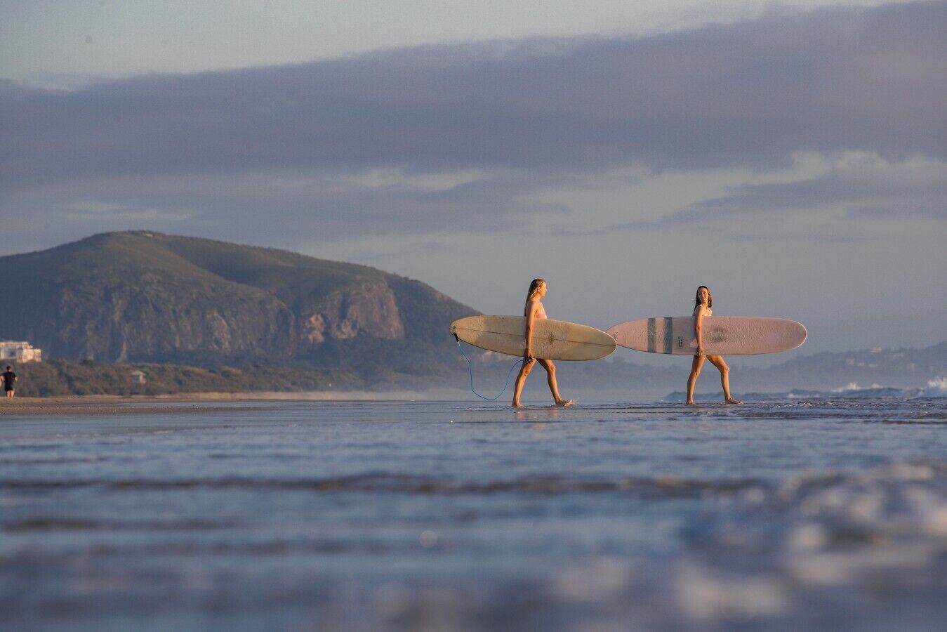 Two people walking on a beach with surfboards, a mountain in the background, and a building on the left.