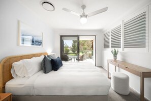 Ein Schlafzimmer mit Bett, Deckenventilator, Fenster mit Jalousien, Nachttisch und Blick ins Freie.