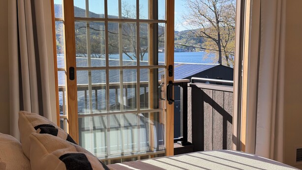 Une chambre avec une grande fenêtre, un lit recouvert d’une couvre-lit à motifs, et une vue sur un lac et des collines.