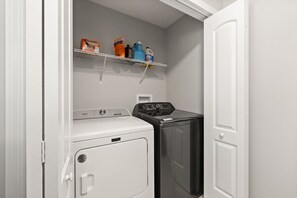 Een wasserette met een witte droger, een zwarte wasmachine en planken met schoonmaakmiddelen.