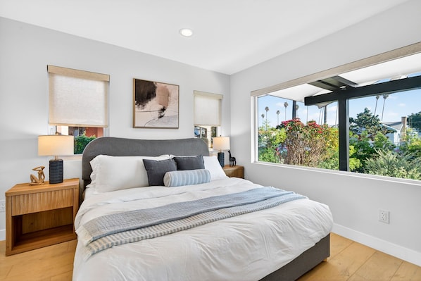 A bedroom with a large bed, two bedside tables, a window with a view of greenery, and a ceiling light.