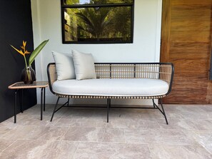 A rattan sofa with cushions, a small table with a plant, and a window with a view of trees.