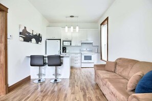 A modern kitchen with white cabinets, a refrigerator, a microwave, and a coffee maker. There are two black bar stools and a beige sofa with a blue pillow.