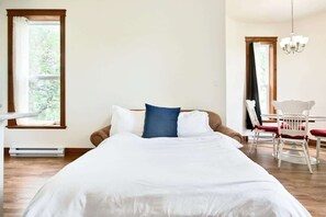 A bedroom with a large bed, a dining table with chairs, and a window with curtains.