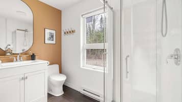 A bathroom with a white vanity, a toilet, a shower with a glass door, and a window with a view of trees.