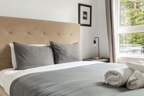 A neatly made bed with a tufted headboard, two pillows, a bedside table with a lamp, and a window with a view of greenery.