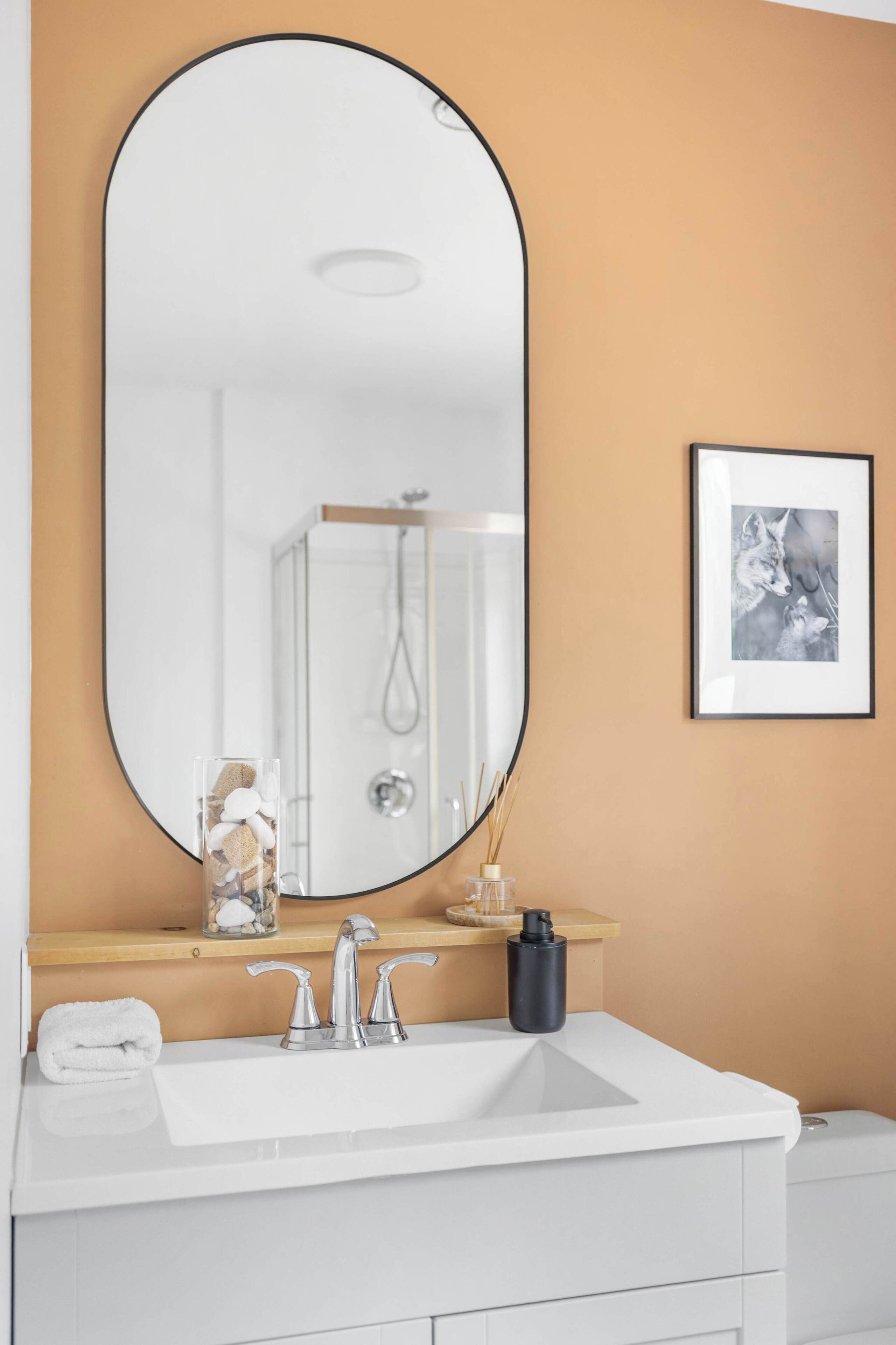 Ein Badezimmer mit einem weißen Waschbecken, einem großen Spiegel, einem gerahmten Bild an der Wand und einer sichtbaren Dusche im Hintergrund.