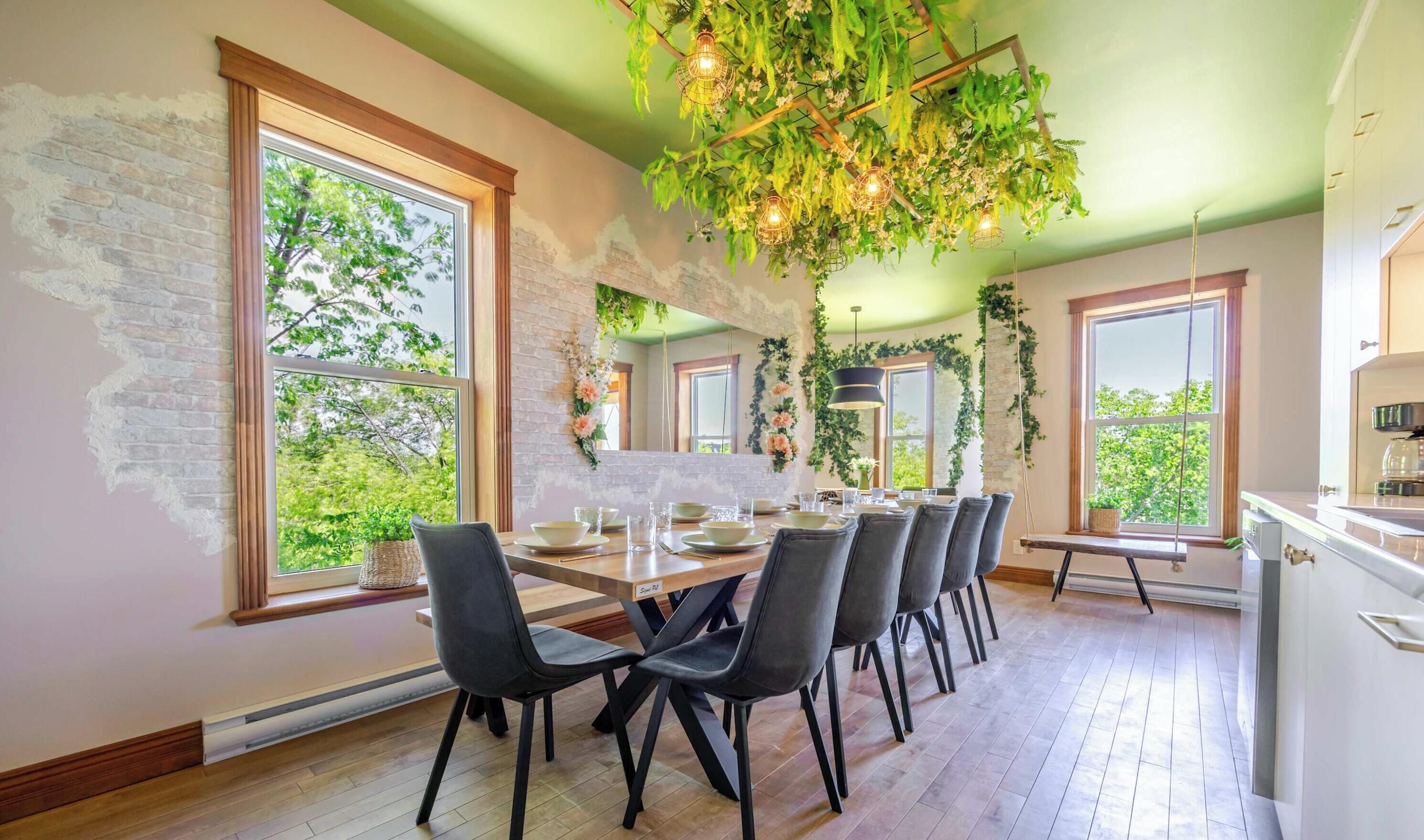 Un espace repas avec un mur en briques, de grandes fenêtres et un plafond orné de plantes suspendues et de luminaires.
