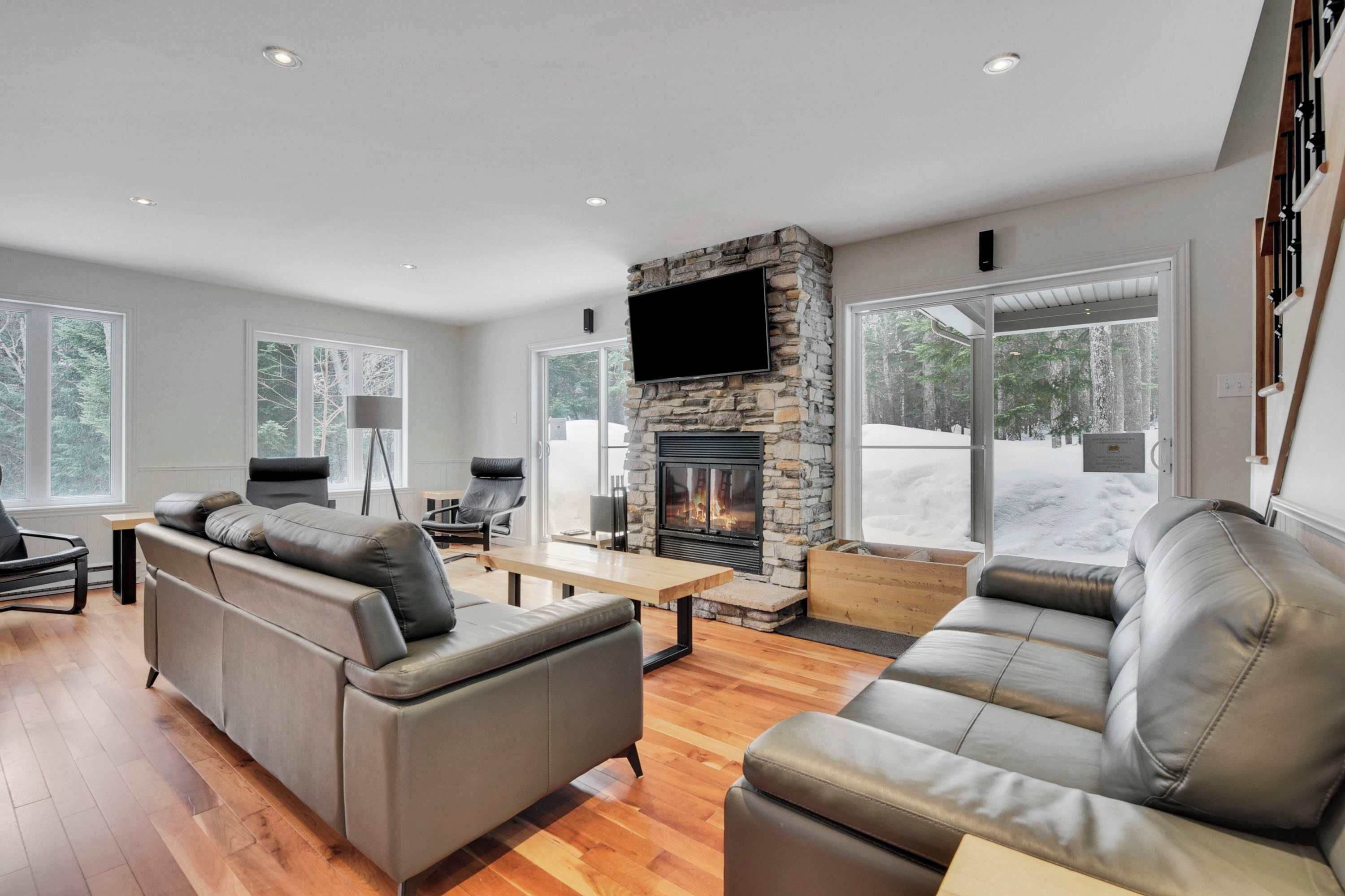 A modern living room with a stone fireplace, leather sofas, a flat-screen TV, and large windows offering a view of the outdoors.