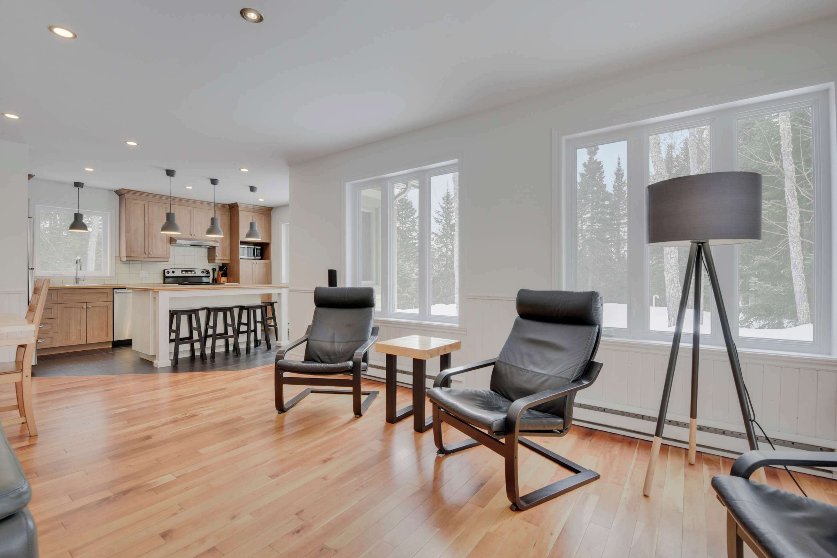 A modern kitchen with wooden cabinets, a central island, and bar stools. There are two black leather chairs and a floor lamp near large windows.