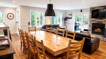 A spacious living room with a large wooden dining table, comfortable seating, a fireplace, and a television mounted on a stone wall.