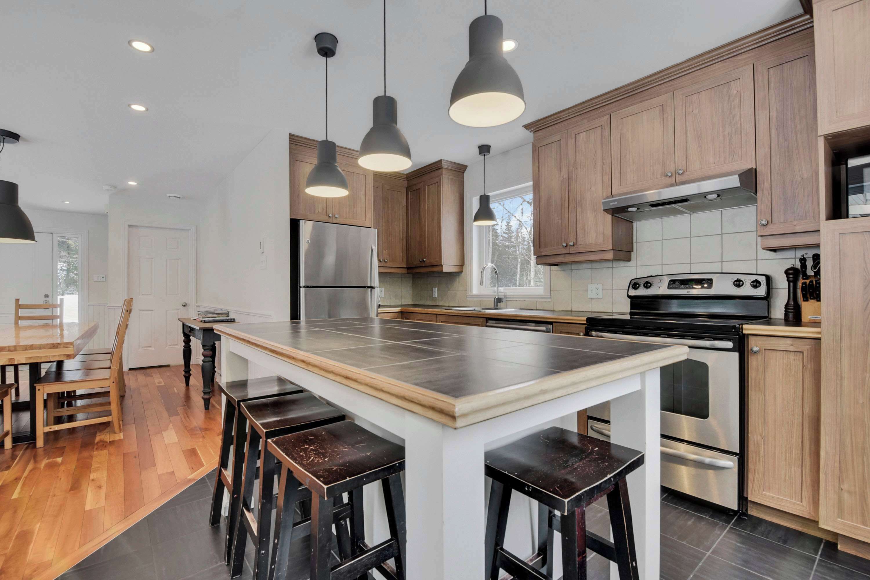 Una cocina moderna con armarios de madera, electrodomésticos de acero inoxidable, una isla central con taburetes, iluminación colgante y vistas al exterior.