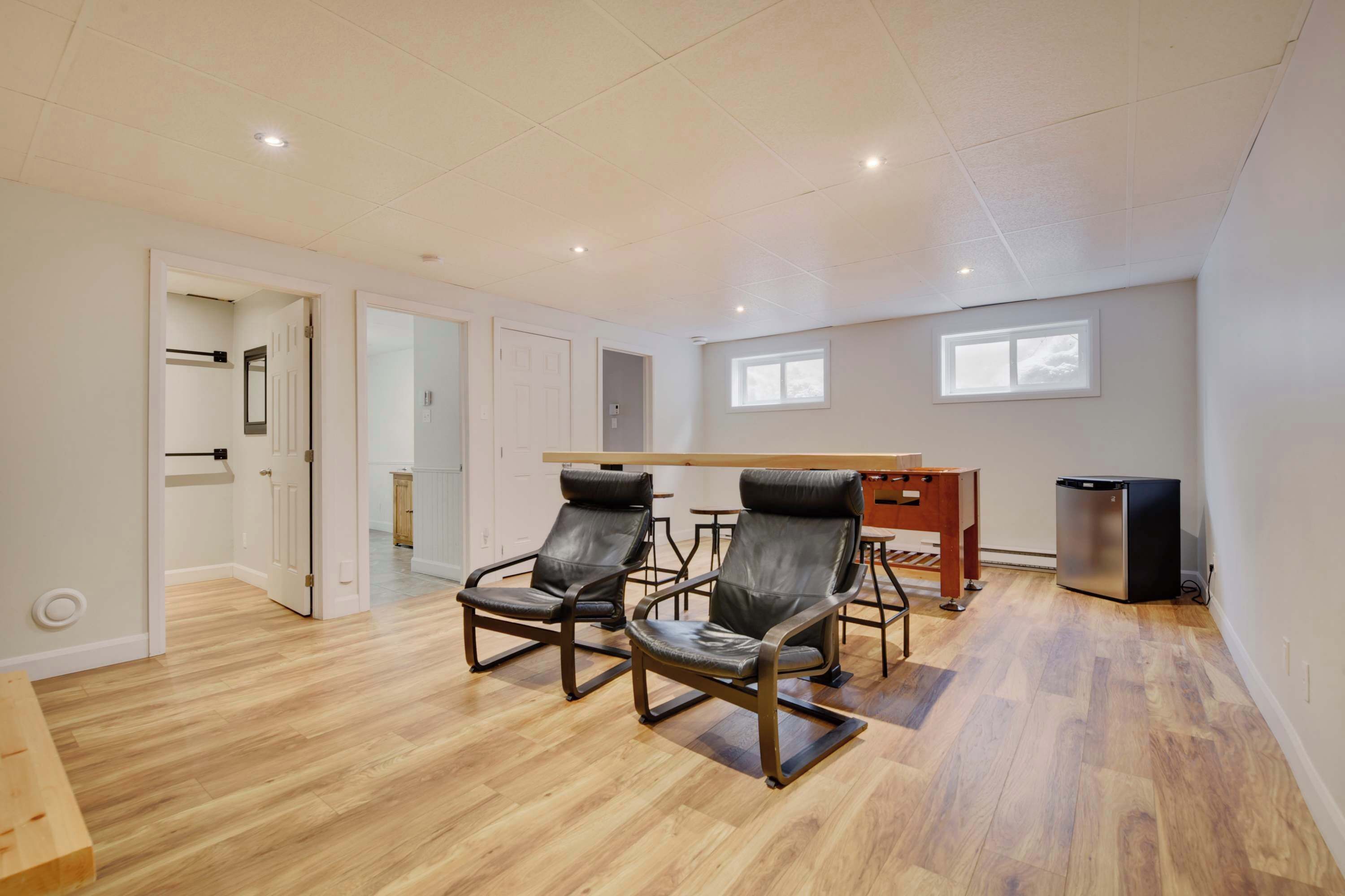 Una habitación amplia con piso de madera, dos sillones de cuero, una mesita, un refrigerador y un ventanal.