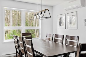 Ein Esszimmer mit Holztisch und Stühlen, großen Fenstern mit Blick auf einen Wald und zwei gerahmten Bildern an der Wand.