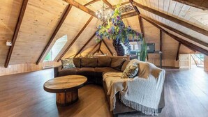 An attic room with a wooden ceiling, a large window, a sofa, a round coffee table, and a decorative hanging plant.