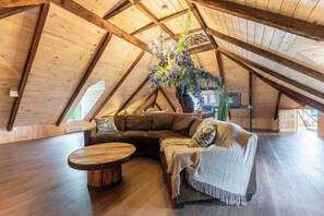 Ein Dachzimmer mit Holzbalkendecke, einem großen Fenster, einer Couch, einem runden Couchtisch und einer dekorativen Hängelpflanze.