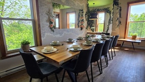 Comedor con una larga mesa de madera dispuesta para seis personas, rodeada de sillas negras, y amplias ventanas que ofrecen vistas a la vegetación exterior.