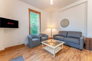 Una sala de estar con una televisión de pantalla plana, dos sillones, un sofá, una mesa de centro de vidrio, una lámpara de pie y un espejo en la pared.