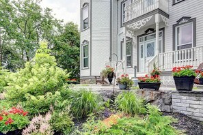 A two-story building with a porch, stone steps, and a garden with various plants and flowers. - Le Juliette  Exceptional site  BBQ & Firepit (Lévis)