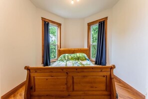 Un dormitorio en esquina con cama de madera, dos ventanas con cortinas y vista a zonas verdes.