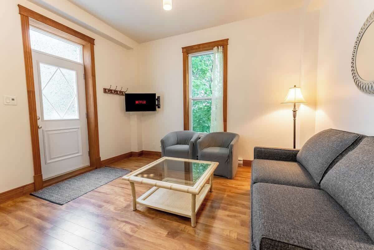 Un salon avec une table basse en verre, deux canapés gris, un téléviseur fixé au mur et une fenêtre avec des rideaux.